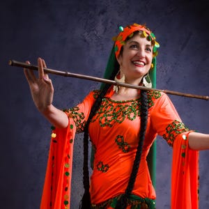 Könnte beinhalten: Ein traditionelles orangefarbenes Kostüm mit grünen und goldenen Akzenten. Das Kostüm hat lange Ärmel, einen fließenden Rock und dekorative Verzierungen. Die Person hält eine Holzflöte und trägt ein Stirnband und Ohrringe.