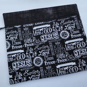 May include: A black zippered pouch featuring a white repeating pattern of religious text and symbols including the words Jesus, Lamb of God, Savior, Lord, King of Kings, Prince of Peace, Emmanuel, and Bread of Life, with a solid black fabric top border.