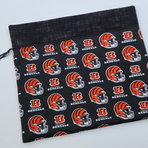 May include: Black zippered pouch featuring a repeating pattern of orange and black Cincinnati Bengals football helmets and logos. The pouch has a black top and a black zipper pull. The text "Cincinnati Bengals" is visible.