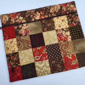 May include: A rectangular zippered pouch featuring a patchwork design of various fabric squares in shades of deep red, dark brown, tan, and cream. The top section has a floral print, and a dark brown zipper with a leather pull is attached.