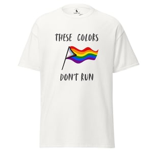 Puede incluir: Camiseta blanca con el texto "THESE COLORS DON'T RUN" encima y debajo de un gráfico de la bandera del arcoíris. La bandera incluye los colores de la bandera del orgullo LGBTQ+ y la bandera del orgullo transgénero.