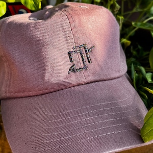 May include: A dusty rose baseball cap with a curved brim. The cap features a stitched design on the front. The cap is made of a soft fabric and has a classic, casual style.
