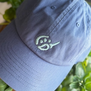 May include: A blue baseball cap with a curved brim. The cap features a light green embroidered design on the front. The cap is made of a soft fabric and has a classic six-panel design.