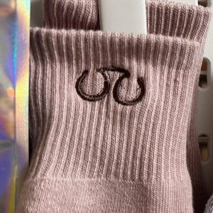 May include: Close-up of a pair of light pink ribbed socks. The socks have a dark brown embroidered design on the cuff. The socks are folded and appear to be new.
