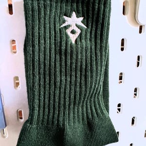 May include: Dark green ribbed knit sock with a white embroidered starburst design. The sock is displayed against a white pegboard background. The cuff is folded over.