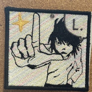 Op de afbeelding: Een vierkante geborduurde patch met een zwart-wit anime-figuur met warrig haar die met de hand een L-vorm maakt. De achtergrond heeft diagonale pastelkleurige regenboogstrepen, een gouden vierpuntige ster en de letter L.