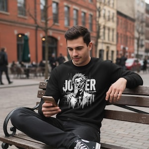 May include: Black sweatshirt featuring a graphic of the Joker with the word "JOKER" in white block letters. The design is a black and white illustration of the Joker's face. The sweatshirt is long-sleeved and made of a soft material.