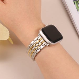 Puede incluir: Una correa de reloj plateada y dorada está sujeta a un reloj inteligente plateado. La correa tiene un diseño tejido con eslabones plateados y dorados alternados. El reloj está en una muñeca.