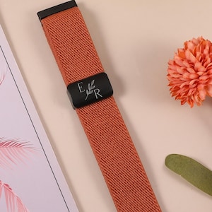 Op de afbeelding: Een oranje horlogeband met zwarte hardware. De band heeft een zwarte gesp met de letters "E" en "R" en een bladontwerp. De band is gemaakt van een geweven materiaal en wordt gepresenteerd op een lichtroze ondergrond.
