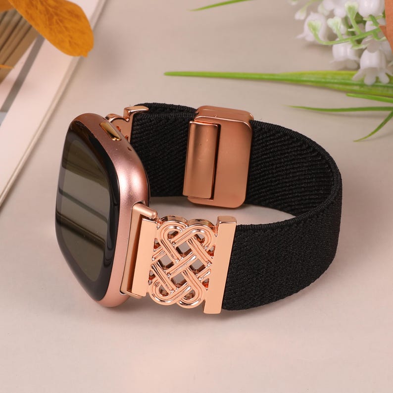 K&ouml;nnte beinhalten: Ros&eacute;goldene Smartwatch mit einem schwarzen elastischen Band. Das Band hat einen dekorativen ros&eacute;goldenen Verschluss und ein dekoratives ros&eacute;goldenes Element mit einem komplizierten Design. Das Zifferblatt ist schwarz.