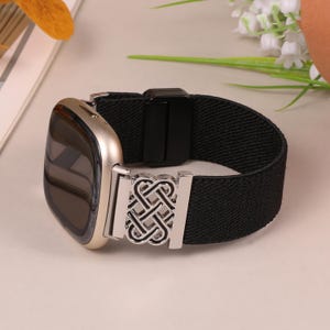 K&ouml;nnte beinhalten: Eine goldfarbene Smartwatch mit einem schwarzen elastischen Armband. Das Armband hat einen silberfarbenen dekorativen Verschluss mit einem komplizierten keltischen Knotenmuster. Die Uhr wird auf einer hellen Oberfl&auml;che pr&auml;sentiert.
