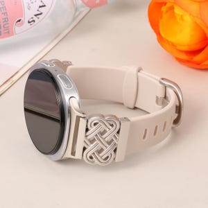 Op de afbeelding: Een smartwatch met een rond zwart scherm en een zilverkleurige metalen behuizing. De horlogeband is gemaakt van glad beige siliconen en heeft een decoratieve zilverkleurige metalen connector met een ingewikkeld, symmetrisch Keltisch knoopmotief.