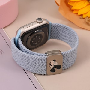 Puede incluir: Un reloj inteligente plateado con una correa tejida azul claro. La correa tiene un cierre plateado con un diseño de Mickey Mouse. El reloj se muestra sobre una superficie clara, con un ramo de flores en el fondo.
