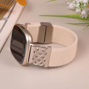 K&ouml;nnte beinhalten: Ein beiges elastisches Uhrenarmband mit einem silberfarbenen Metallverschluss und einem dekorativen Knotendesign. Das Zifferblatt der Uhr ist silberfarben mit einem schwarzen Bildschirm. Das Armband besteht aus gewebtem Material.