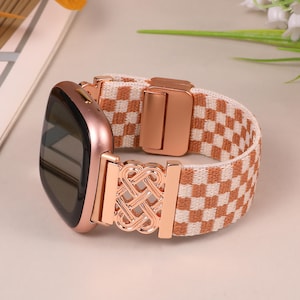 K&ouml;nnte beinhalten: Eine ros&eacute;goldene Smartwatch mit einem karierten, beige-wei&szlig;en Armband. Das Armband hat einen ros&eacute;goldenen Verschluss und dekorative Elemente. Das Zifferblatt der Uhr ist schwarz und das Armband besteht aus gewebtem Material.