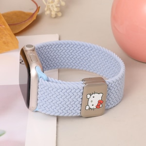 Puede incluir: Una correa de reloj elástica trenzada de color azul claro unida a una caja de reloj inteligente plateada. La correa cuenta con un dije deslizante metálico rectangular decorado con un gráfico blanco de Hello Kitty, con un lazo rojo y detalles negros.