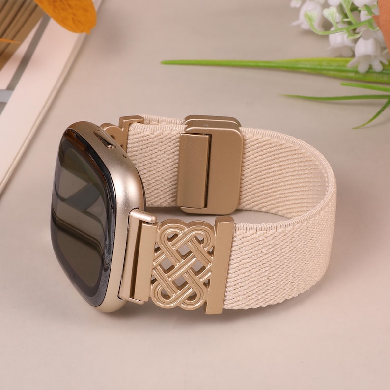 K&ouml;nnte beinhalten: Eine goldfarbene Smartwatch mit einem cremefarbenen elastischen Armband. Das Armband hat einen goldfarbenen dekorativen Verschluss mit einem komplizierten Knotendesign. Das Zifferblatt der Uhr ist schwarz und das Armband ist etwa 2,5 cm breit.
