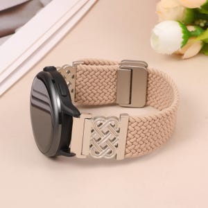 May include: A smartwatch with a round black face and a beige braided elastic watch band. The band features silver-toned metal connectors with an intricate openwork knot design and a rectangular silver-toned metal sliding adjustment buckle.