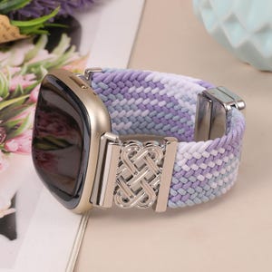 Puede incluir: Un smartwatch con caja dorada y correa trenzada en tonos morado, blanco y gris. La correa presenta un cierre plateado y detalles metálicos decorativos. Un accesorio moderno y funcional para el día a día.