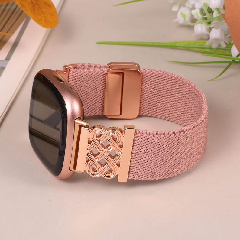 K&ouml;nnte beinhalten: Eine ros&eacute;goldene Smartwatch mit passendem rosa elastischem Armband. Das Armband hat einen ros&eacute;goldenen Verschluss und eine dekorative Schnalle. Die Uhr wird auf einer hellrosa Oberfl&auml;che mit floralen Akzenten pr&auml;sentiert.