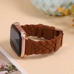 Puede incluir: Una correa de reloj trenzada de cuero marrón con una esfera de reloj de oro rosa. La correa está hecha de cuero tejido y tiene una hebilla dorada. La esfera del reloj es rectangular con una pantalla negra.