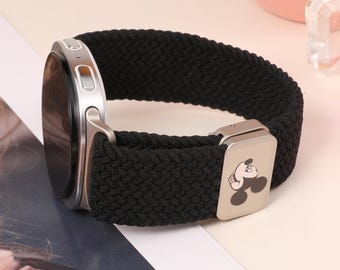 Könnte beinhalten: Ein schwarzes, geflochtenes elastisches Uhrenarmband an einem silberfarbenen Smartwatch-Gehäuse. Das Armband verfügt über einen rechteckigen silbernen Metallschieber, der mit einer kleinen, aufgedruckten Grafik von Mickys Gesicht verziert ist.