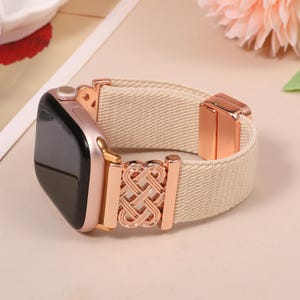 Pode incluir: Smartwatch em ouro rosa com uma pulseira elástica bege. A pulseira apresenta um fecho decorativo em ouro rosa e uma textura tecida. O mostrador do relógio é preto. A pulseira é projetada para esticar.