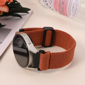 May include: A silver smartwatch with a round black screen and a rust-colored elastic band. The band has a black buckle. The watch is resting on a white surface.