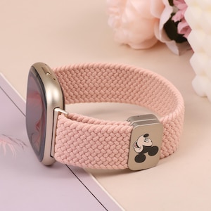 May include: A light pink watch band with a silver watch face. The band is made of a woven material. The clasp features a black and white Mickey Mouse design.