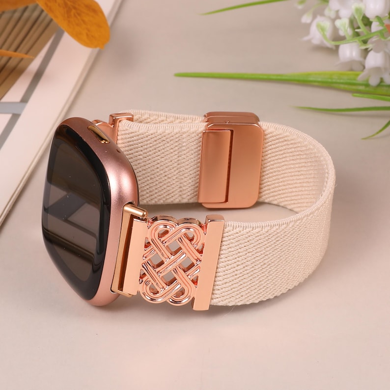 K&ouml;nnte beinhalten: Eine ros&eacute;goldene Smartwatch mit einem cremefarbenen elastischen Band. Das Band hat einen dekorativen ros&eacute;goldenen Verschluss und eine gewebte Textur. Das Zifferblatt der Uhr ist schwarz. Das Band ist etwa 1,9 cm breit.