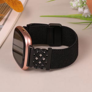 K&ouml;nnte beinhalten: Eine ros&eacute;goldene Smartwatch mit einem schwarzen elastischen Armband. Das Armband hat einen dekorativen schwarzen Metallverschluss mit einem komplizierten Knotendesign. Die Uhr wird auf einer hellen Oberfl&auml;che pr&auml;sentiert.