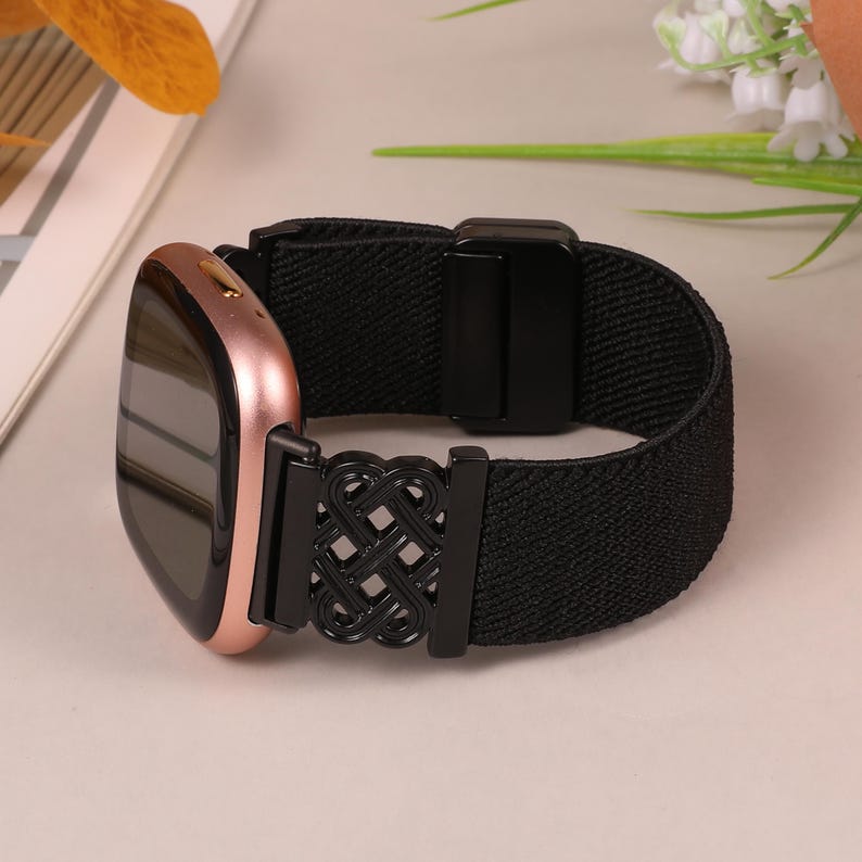 K&ouml;nnte beinhalten: Eine ros&eacute;goldene Smartwatch mit einem schwarzen elastischen Armband. Das Armband hat einen dekorativen schwarzen Metallverschluss mit einem komplizierten Knotendesign. Die Uhr wird auf einer hellen Oberfl&auml;che pr&auml;sentiert.