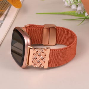K&ouml;nnte beinhalten: Eine ros&eacute;goldene Smartwatch mit einem terracottafarbenen elastischen Armband. Das Armband hat einen ros&eacute;goldenen dekorativen Verschluss mit einem komplizierten Knotendesign. Das Zifferblatt der Uhr ist schwarz.
