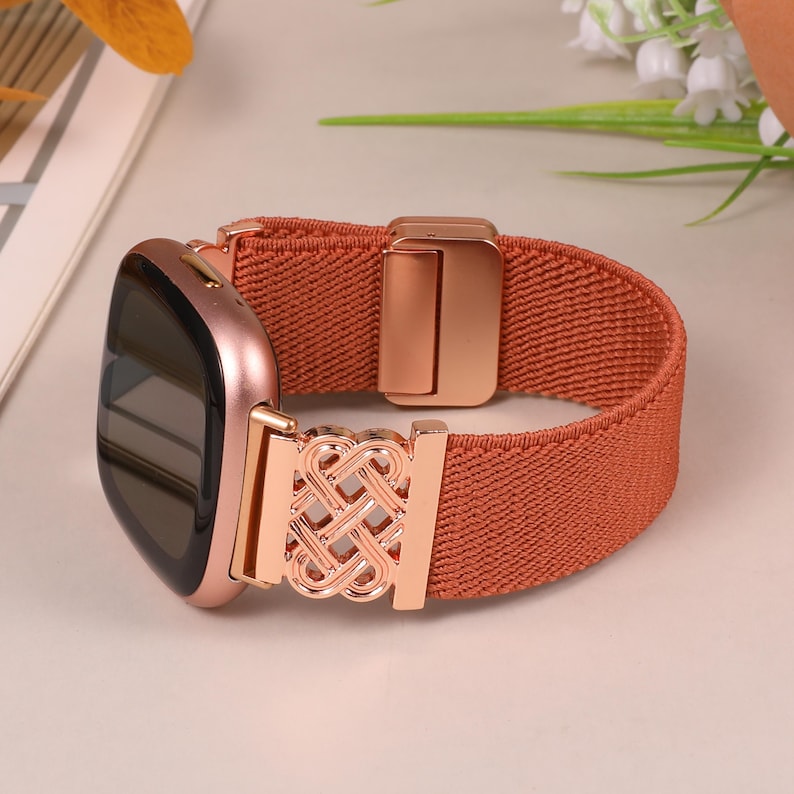 K&ouml;nnte beinhalten: Eine ros&eacute;goldene Smartwatch mit einem terracottafarbenen elastischen Armband. Das Armband hat einen ros&eacute;goldenen dekorativen Verschluss mit einem komplizierten Knotendesign. Das Zifferblatt der Uhr ist schwarz.