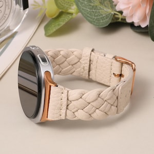 Puede incluir: Un reloj inteligente con pantalla redonda negra, caja de metal plateado y oro rosa, y una correa de cuero trenzado color crema. La correa cuenta con una hebilla de metal en oro rosa y un acabado texturizado, unida a la esfera por soportes.