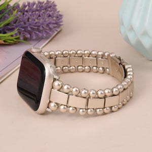May include: Silver-toned smartwatch with a matching beaded band. The band features a combination of rectangular links and small, round beads. The watch face is black with a silver frame. The watch is displayed on a light-colored surface.