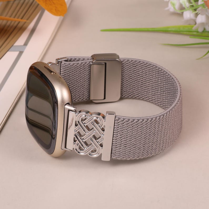K&ouml;nnte beinhalten: Ein graues elastisches Uhrenarmband mit einem silberfarbenen Metallverschluss und dekorativem Akzent. Das Zifferblatt der Uhr ist goldfarben mit einem schwarzen Bildschirm. Das Armband ist dehnbar und passt sich bequem dem Handgelenk an.