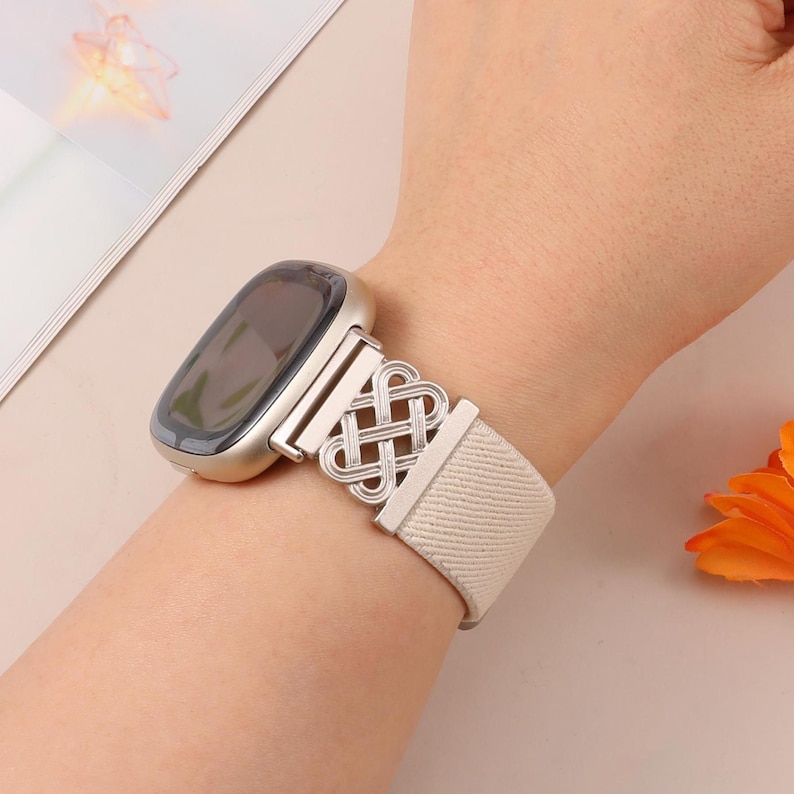 K&ouml;nnte beinhalten: Eine silberne Smartwatch mit einem hellbeigen Armband. Das Armband hat ein dekoratives silbernes Knotendesign. Die Uhr wird am Handgelenk getragen, mit einem unscharfen Hintergrund.