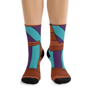 May include: A pair of crew-length socks featuring a geometric pattern of brown, purple, and turquoise shapes. The design consists of angular blocks and stripes arranged in a repeating motif, finished with solid black toe and heel sections.