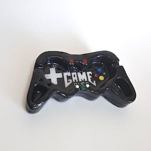Könnte beinhalten: Schwarzer Aschenbecher in Form eines Videospiel-Controllers. Der Aschenbecher hat ein weißes Kreuz und die Worte "GAME OVER" in Weiß. Er verfügt über rote, blaue, grüne und gelbe Knöpfe sowie zwei Aschenbecherablagen.