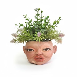 May include: A unique, hand-painted ceramic planter shaped like a face, with detailed eyes, lips, and ears. The planter is filled with a variety of green and purple faux plants. The face has a light skin tone with darker shading.