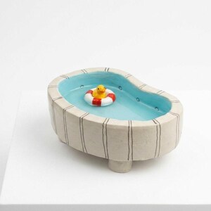 May include: A ceramic bowl shaped like a swimming pool with light blue interior and off-white exterior featuring vertical black stripes. A small yellow rubber duck sits inside a red and white striped life preserver ring floating in the centre.