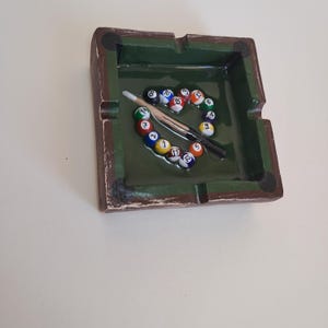 May include: A rectangular ashtray with a green interior and a brown, textured exterior. The ashtray is decorated with a miniature pool table design, featuring colorful billiard balls and a cue stick. The balls are arranged in a circular pattern.