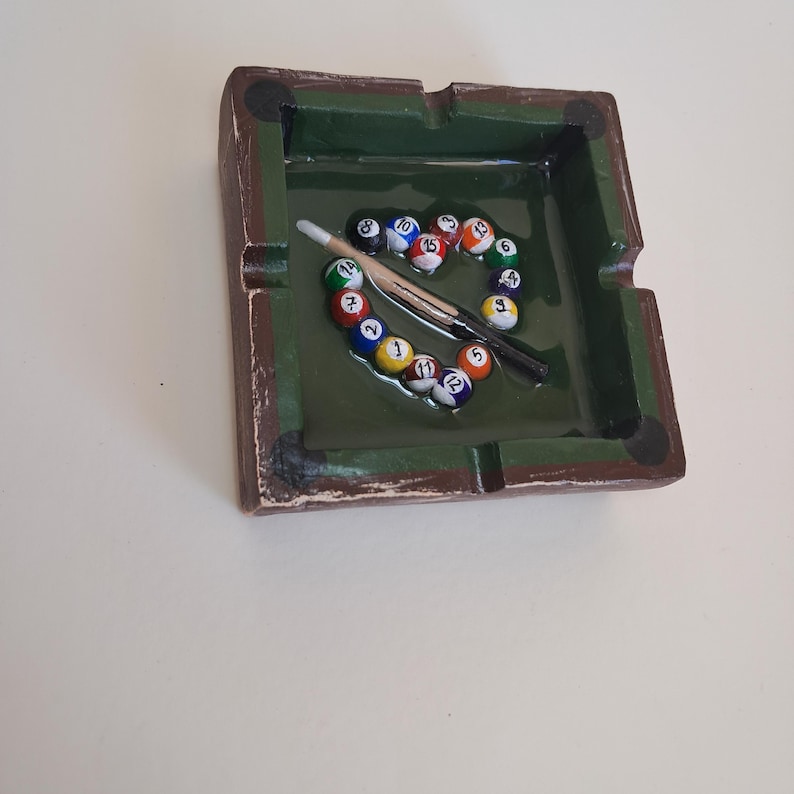 May include: A rectangular ashtray with a green interior and a brown, textured exterior. The ashtray is decorated with a miniature pool table design, featuring colorful billiard balls and a cue stick. The balls are arranged in a circular pattern.
