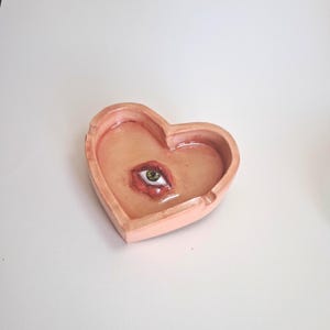 May include: A heart-shaped ashtray in a peach color, featuring a painted eye design. The eye is green with red accents around it. The ashtray is made of ceramic or a similar material.