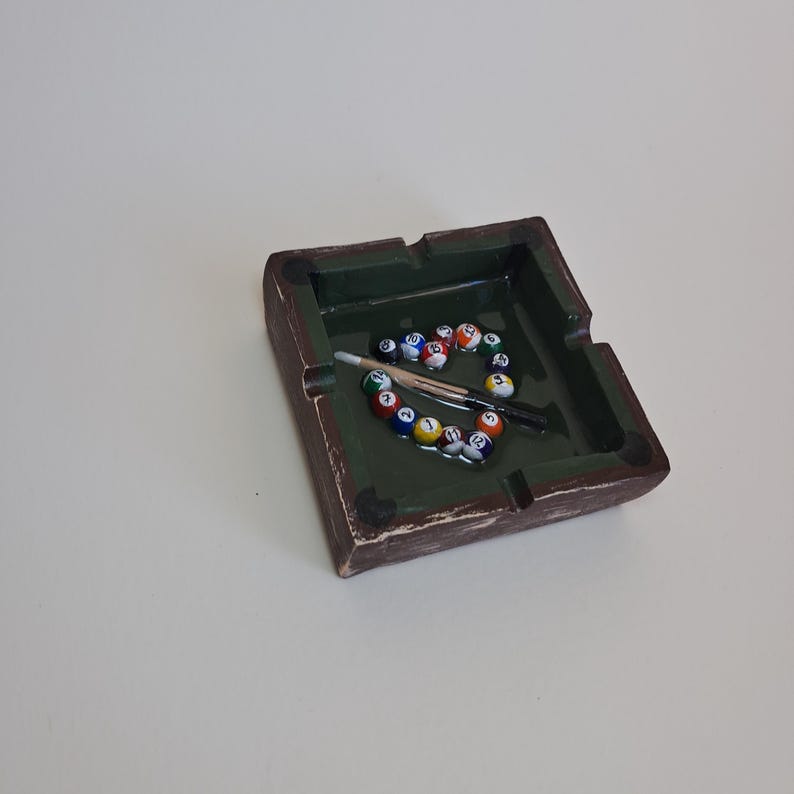 May include: A square ashtray with a green interior and brown exterior, resembling a miniature pool table. Inside, a circle of miniature billiard balls surrounds a small pool cue and a black cue stick. The ashtray is on a white surface.