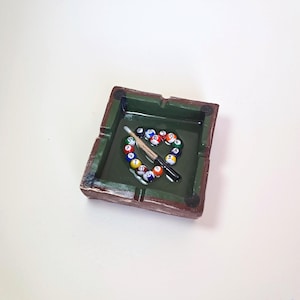May include: A square ashtray with a green interior and a brown exterior. Inside, a heart shape is formed by colorful billiard balls, with a cue stick resting across them. The ashtray has a slot for cigarettes.