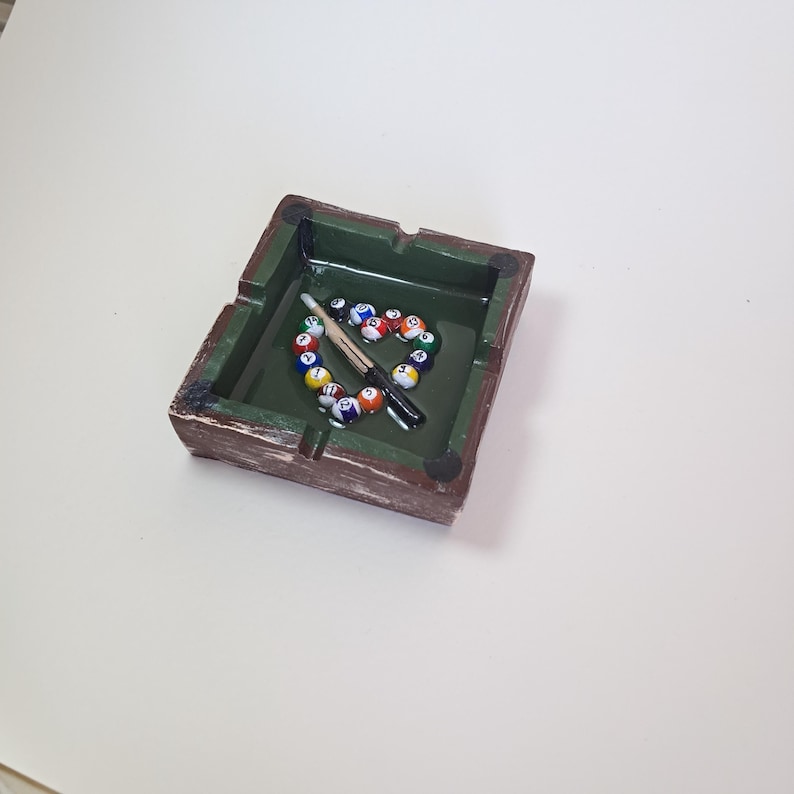 May include: A square ashtray with a green interior and a brown, textured exterior. Inside, a heart shape is formed by colorful billiard balls, with a cue stick resting across them. The ashtray has small, round feet.