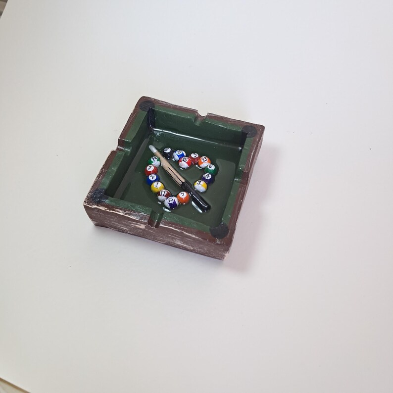 May include: A square ashtray with a green interior and a brown, textured exterior. Inside, a heart shape is formed by colorful billiard balls, with a cue stick resting across them. The ashtray has small, round feet.