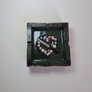 May include: A square, green ashtray with a brown rim. Inside, a heart shape is formed by colorful billiard balls and a cue stick. The ashtray has a glossy finish and is designed to resemble a pool table.